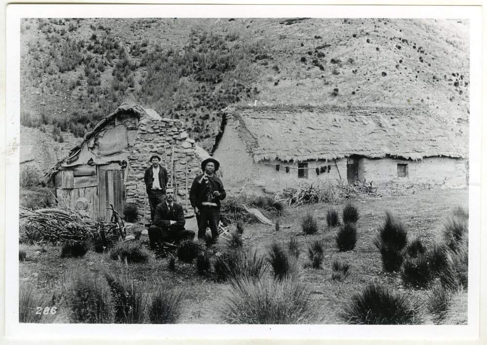 Reverend George McNeur sits between two Chinese men, behind them is a stone and clay brick dwelling