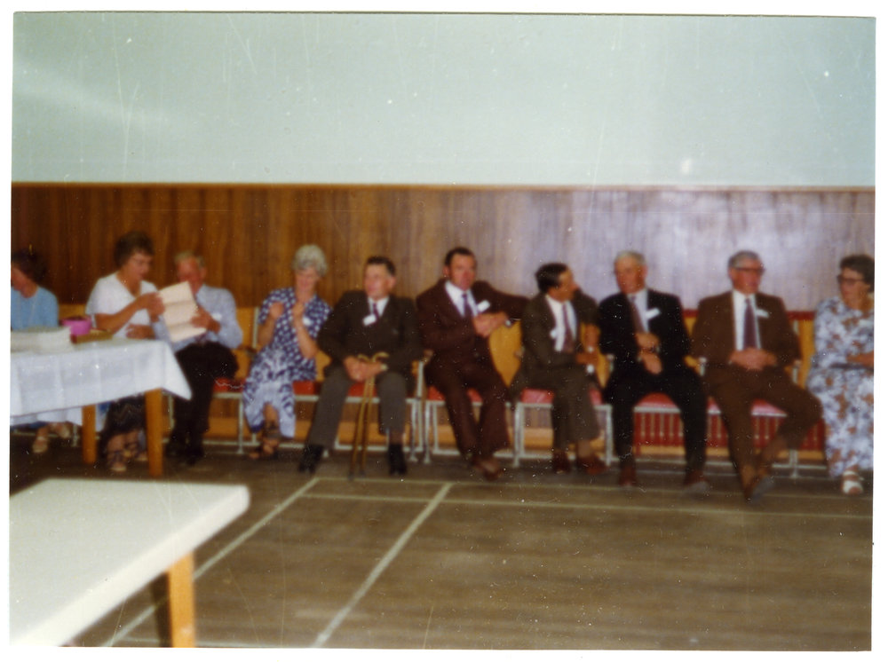 Group of men and women, who belong to Cromwell Church, are seated along the wall of a sports gymnasium 