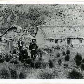 Reverend George McNeur sits between two Chinese men, behind them is a stone and clay brick dwelling