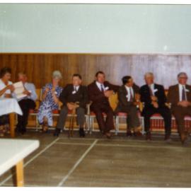 Group of men and women, who belong to Cromwell Church, are seated along the wall of a sports gymnasium 