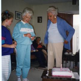 Alicia Forbes, Margaret Weir and Hilda Muff are standing in a lounge, about to light a candle on a celebration cake
