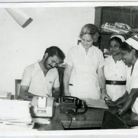 Beryl Howie stands over the shoulder of an Indian orderly, whilst two unidentified Indian nurses look on
